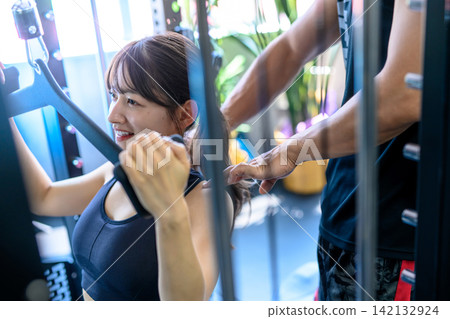 一名女子正在健身房使用下拉式器材鍛煉,並接受教練的指導。 一名女子正在健身房使用下拉式器材鍛煉,並接受教練的指導。 142132924