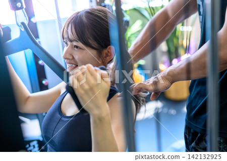 A woman working out with a lat pulldown machine, receiving instructions from a trainer at the gym. Art Trimming Up A woman working out with a lat pulldown machine, receiving instructions from a trainer at the gym. Art Trimming Up 142132925