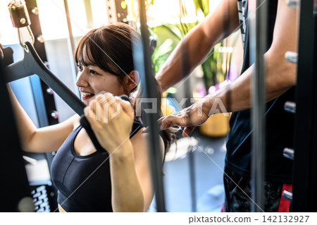 一位女士正在使用下拉器材運動;在健身房接受教練的指導;白天的彩色藝術作品。 一位女士正在使用下拉器材運動;在健身房接受教練的指導;白天的彩色藝術作品。 142132927