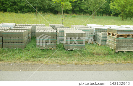Concrete paving blocks stacked interlocking pavers rectangular, bricks curb blocks stacked wooden pallets green plastic banding straps strapping, stored outdoors grassy verge and a paved road stored Concrete paving blocks stacked interlocking pavers rectangular, bricks curb blocks stacked wooden pallets green plastic banding straps strapping, stored outdoors grassy verge and a paved road stored 142132943