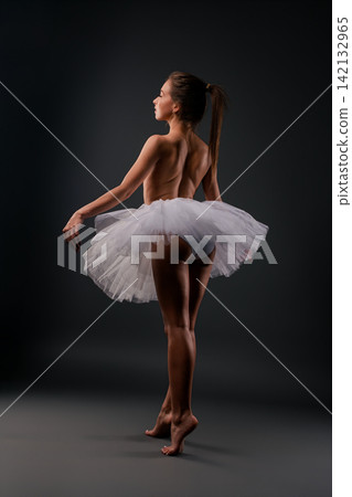 Ballerina in white tutu posing barefoot against dark studio background Ballerina in white tutu posing barefoot against dark studio background 142132965