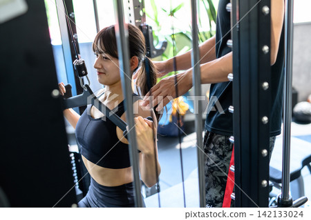 A woman works out using a lat pulldown machine, receiving instructions from a trainer at the gym. A woman works out using a lat pulldown machine, receiving instructions from a trainer at the gym. 142133024