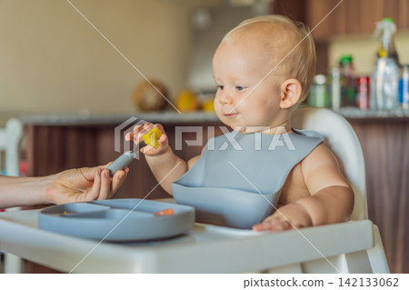 Infant practices self feeding with vegetables in a kitchen, showing baby led weaning, early nutrition and family care concept Infant practices self feeding with vegetables in a kitchen, showing baby led weaning, early nutrition and family care concept 142133062