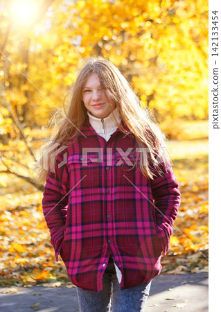 Girl walks through a park with autumn leaves on the ground during a sunny day in a bright plaid jacket Girl walks through a park with autumn leaves on the ground during a sunny day in a bright plaid jacket 142133454