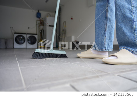 Woman sweeps dust and debris from tiled garage floor with a broom Woman sweeps dust and debris from tiled garage floor with a broom 142133563