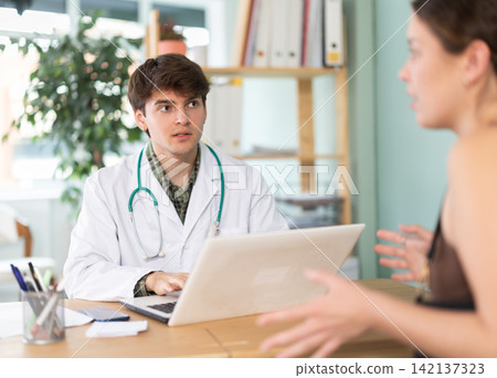 Experienced young male doctor wearing uniform with stethoscope doing check up of sick and health young girl patient sitting with laptop at table in medical office at hospital Experienced young male doctor wearing uniform with stethoscope doing check up of sick and health young girl patient sitting with laptop at table in medical office at hospital 142137323