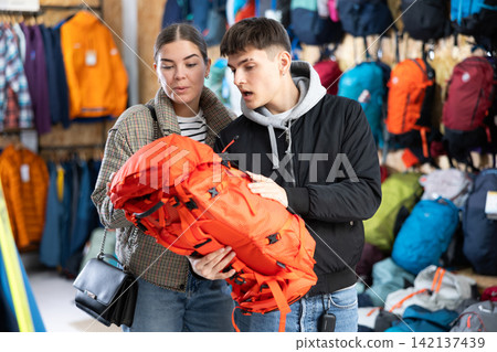 Young married couple in winter clothes chooses a durable backpack for a winter hike in tourist equipment store Young married couple in winter clothes chooses a durable backpack for a winter hike in tourist equipment store 142137439