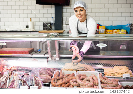 Young female butcher arranging raw sausages in glass counter Young female butcher arranging raw sausages in glass counter 142137442