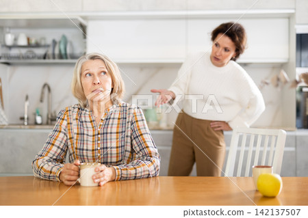 Upset old woman sitting at the kitchen table with her back to woman quarreling to her Upset old woman sitting at the kitchen table with her back to woman quarreling to her 142137507