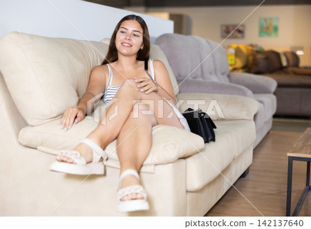 In furniture store,young girl is sitting on sofa with ottoman In furniture store,young girl is sitting on sofa with ottoman 142137640