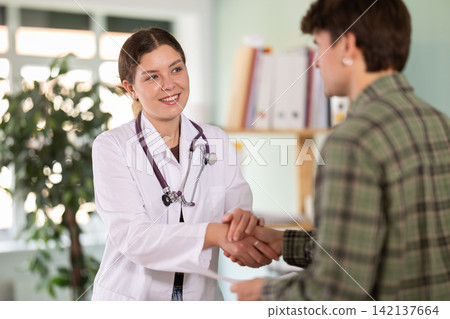 Smiling young female doctor or nurse wearing white uniform with stethoscope shaking hands with young guy patient congratulating on good test results or recovery from illness in medical office at Smiling young female doctor or nurse wearing white uniform with stethoscope shaking hands with young guy patient congratulating on good test results or recovery from illness in medical office at 142137664