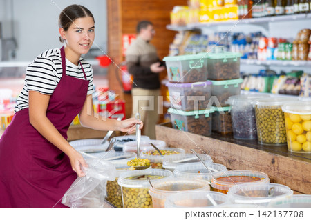 Smiling young vendoress pouring marinated olives into bag in grocery store Smiling young vendoress pouring marinated olives into bag in grocery store 142137708