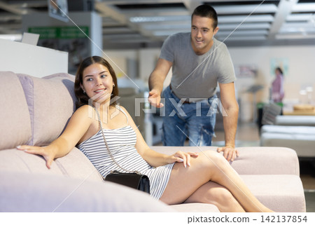 Couple chooses furniture in store.Wife is sitting on sofa,husband is standing next to her Couple chooses furniture in store.Wife is sitting on sofa,husband is standing next to her 142137854