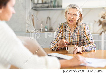 Positive old woman sitting at the kitchen-table face to face with middle-aged female agent Positive old woman sitting at the kitchen-table face to face with middle-aged female agent 142137858