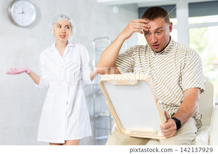 Unsatisfied middle-aged man looking in the mirror sitting on clinical chair Unsatisfied middle-aged man looking in the mirror sitting on clinical chair 142137929