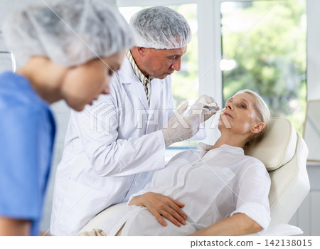 Man doctor perform rejuvenating injection in area around mouth to mature woman, female nurse assists Man doctor perform rejuvenating injection in area around mouth to mature woman, female nurse assists 142138015
