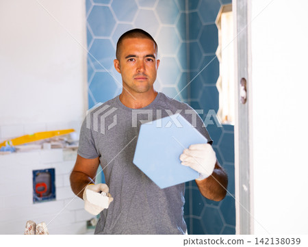 Skilled man builder in ordinary clothes posing with ceramic tiles while repair and decoration in apartment Skilled man builder in ordinary clothes posing with ceramic tiles while repair and decoration in apartment 142138039