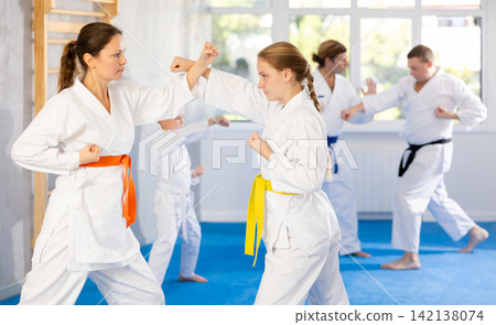 Young girl and woman in kimono and colored belt practicing karate punch block during group martial arts lesson in gym Young girl and woman in kimono and colored belt practicing karate punch block during group martial arts lesson in gym 142138074
