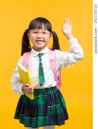 A cute schoolgirl wearing glasses waves her hand in a greeting or answering a question in class against a bright yellow background. A cute schoolgirl wearing glasses waves her hand in a greeting or answering a question in class against a bright yellow background. 142138725