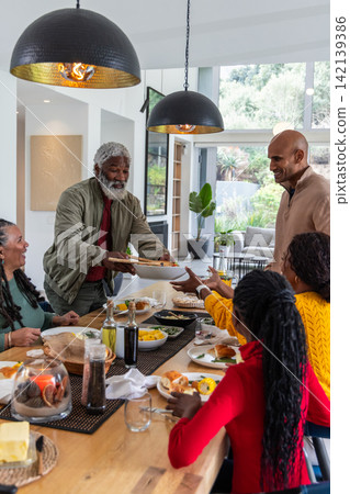 African American family with senior man passing bowl at dining table, wearing casual sweaters African American family with senior man passing bowl at dining table, wearing casual sweaters 142139386
