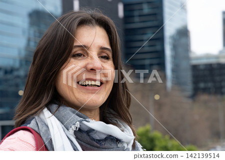 Middle-aged woman smiling, standing on sidewalk with gray scarf, pink top, red strap, glass towers Middle-aged woman smiling, standing on sidewalk with gray scarf, pink top, red strap, glass towers 142139514