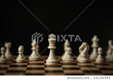 white chess king standing tall on a board, surrounded by blurred pieces on black background white chess king standing tall on a board, surrounded by blurred pieces on black background 142142845