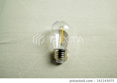 A small clear LED filament light bulb lying on a textured sandy background, representing traveling idea and minimal design. Summer, natural or vacation background 142143075