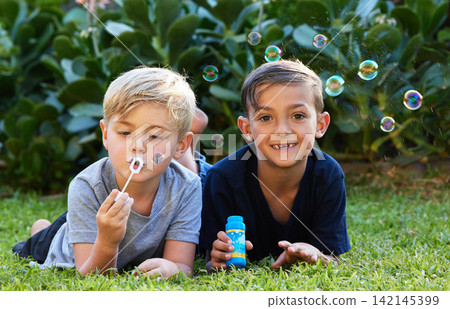 Friends, playing and children blowing bubbles in park for weekend activity, bonding or having fun together. Happy, relax and young boys lying on lawn for childhood, leisure or enjoyment in backyard 142145399