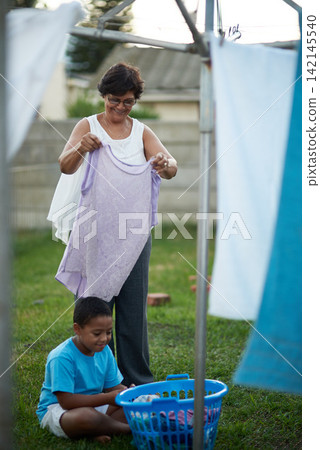 Outdoor, grandmother and grandchild with laundry, help and chore with summer and bonding together. Family, senior woman and boy with clean clothes and activity in backyard with washing and home Outdoor, grandmother and grandchild with laundry, help and chore with summer and bonding together. Family, senior woman and boy with clean clothes and activity in backyard with washing and home 142145540