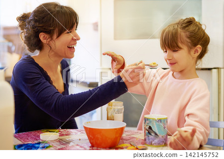 Happy mother, child and feeding with cereal for fiber, vitamin or breakfast in kitchen together at home. Mom, little girl or young daughter with spoon of porridge for healthy meal, nutrition or snack 142145752