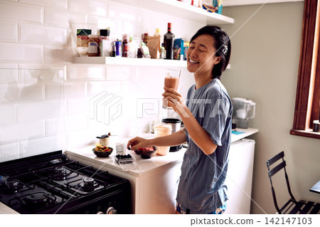 Asian woman, happy and smoothie in kitchen for healthy breakfast, morning routine and body detox in pajamas. Person, smile and drinking organic drink for wellness and vegan diet by counter in home Asian woman, happy and smoothie in kitchen for healthy breakfast, morning routine and body detox in pajamas. Person, smile and drinking organic drink for wellness and vegan diet by counter in home 142147103
