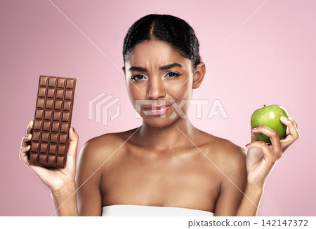 Black woman, health and nutrition with food choice fro balanced diet isolated on pink background. Happy, female person or model and apple or chocolate option for weight loss fruit in studio. Black woman, health and nutrition with food choice fro balanced diet isolated on pink background. Happy, female person or model and apple or chocolate option for weight loss fruit in studio. 142147372