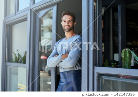 Door, smile and portrait of barista by coffee shop with positive, happy and confident attitude. Crossed arms, pride and man waiter standing by cafe or restaurant entrance for welcome in hospitality. Door, smile and portrait of barista by coffee shop with positive, happy and confident attitude. Crossed arms, pride and man waiter standing by cafe or restaurant entrance for welcome in hospitality. 142147386