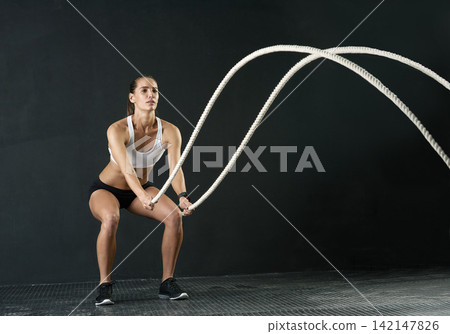 Woman, studio and training with battle rope for fitness, calorie and body workout for vitality or wellness. Girl, squat and exercise on black background for strength, power and endurance or gym Woman, studio and training with battle rope for fitness, calorie and body workout for vitality or wellness. Girl, squat and exercise on black background for strength, power and endurance or gym 142147826