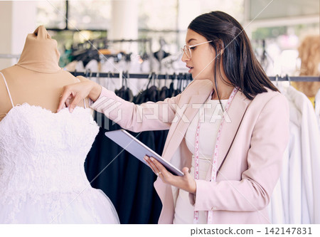 Tablet, wedding dress and fashion designer woman in studio for boutique management or planning. Review, style or textile and seamstress with tape measure in creative workshop to complete online order Tablet, wedding dress and fashion designer woman in studio for boutique management or planning. Review, style or textile and seamstress with tape measure in creative workshop to complete online order 142147831