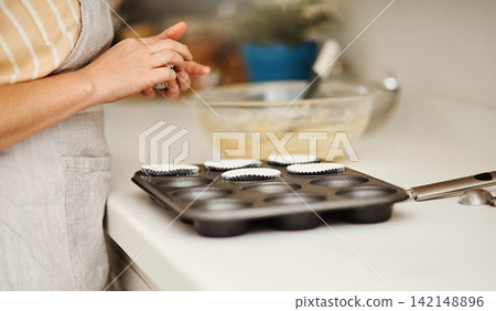 Baker hands, baking tray and cupcakes prepare in kitchen in house for relax or therapeutic hobby. Dessert, cooking and professional person with homemade for small business or home recipe with paper. Baker hands, baking tray and cupcakes prepare in kitchen in house for relax or therapeutic hobby. Dessert, cooking and professional person with homemade for small business or home recipe with paper. 142148896