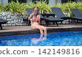 Teenage girl in pink two-piece swimsuit sitting on pool edge with legs in water giving thumbs up to camera while drinking a strawberry cocktail. Happy summer concept 142149816