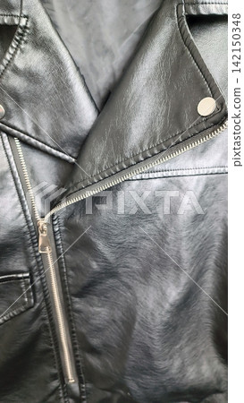 Close up of the upper left side of a black faux leather jacket, showing the lapel, diagonal silver zipper, button, seams, and stitching, with a slightly wrinkled textured surface that reflects light Close up of the upper left side of a black faux leather jacket, showing the lapel, diagonal silver zipper, button, seams, and stitching, with a slightly wrinkled textured surface that reflects light 142150348