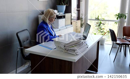 Focused businesswoman working with documents and laptop at her desk in a contemporary office space with natural lighting Focused businesswoman working with documents and laptop at her desk in a contemporary office space with natural lighting 142150635