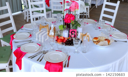 White draped banquet table featuring floral centerpiece, fresh fruits, glowing candles, fuchsia napkins, elegantly prepared for intimate wedding reception dining White draped banquet table featuring floral centerpiece, fresh fruits, glowing candles, fuchsia napkins, elegantly prepared for intimate wedding reception dining 142150683