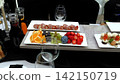 Elegant white ceramic platter presenting rolled meat, citrus, berries, stone fruits, canapes alongside stemware and wine decanters resting on neutral gray surface 142150719