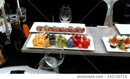Elegant white ceramic platter presenting rolled meat, citrus, berries, stone fruits, canapes alongside stemware and wine decanters resting on neutral gray surface 142150719