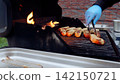 Professional chef wearing protective gloves grilling savory sausages on sizzling barbecue, preparing flavorful meal during sunny outdoor dining experience 142150721