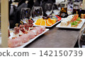 Gourmet catering platters displaying diverse appetizers including grilled meat skewers, fresh fruit selections, and delicate dessert rolls, accompanied by sparkling wine glasses 142150739