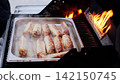 Grilling juicy sausages over gas flames in metal tray, creating smoky barbecue atmosphere during summer outdoor cooking session 142150745