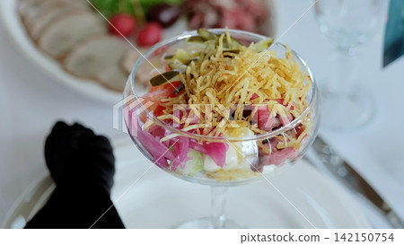 Russian salad - olivier - in glass, garnished with crispy potato straws, served during catering event with charcuterie plate blurring background Russian salad - olivier - in glass, garnished with crispy potato straws, served during catering event with charcuterie plate blurring background 142150754