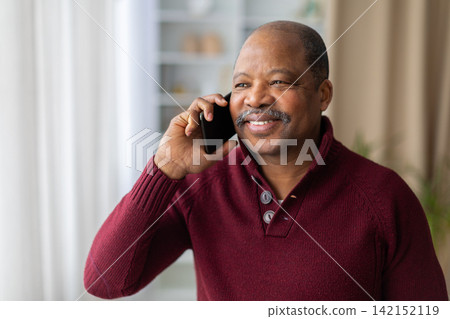 Smiling mature Black man talking on smartphone at home, enjoying phone conversation Smiling mature Black man talking on smartphone at home, enjoying phone conversation 142152119