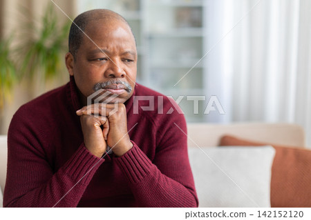 Thoughtful Black man sitting at home with chin resting on clasped hands Thoughtful Black man sitting at home with chin resting on clasped hands 142152120