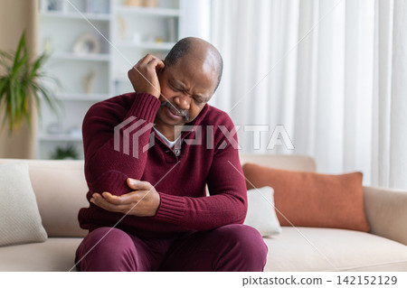 Mature black man rubbing sore elbow in discomfort while sitting on sofa Mature black man rubbing sore elbow in discomfort while sitting on sofa 142152129