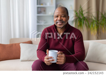 Mature Black man sitting on sofa at home and holding mug Mature Black man sitting on sofa at home and holding mug 142152143